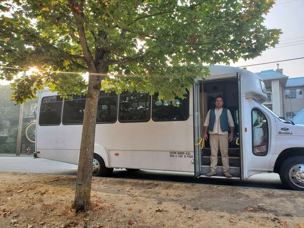 Jorge standing in the doorway of the Culture Caravan bus, parked under a maple tree.