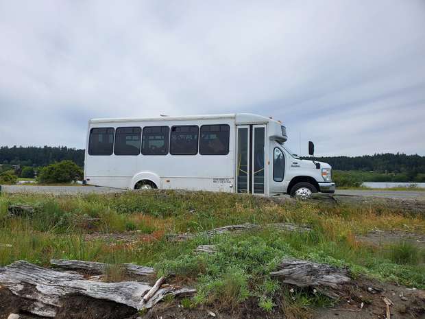 The Culture Caravan bus parked roadside by driftwood, with a lake and forest behind it.